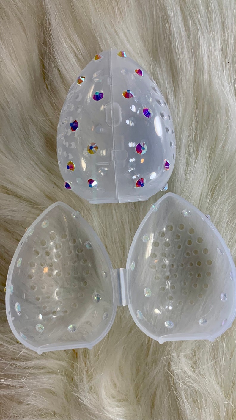 May include: A clear plastic makeup sponge holder with a decorative design of iridescent rhinestones. The holder is divided into two halves, each with a honeycomb-like pattern.