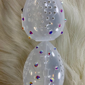 May include: Two clear plastic egg-shaped makeup sponge holders with iridescent rhinestones. The holders are stacked on top of each other.