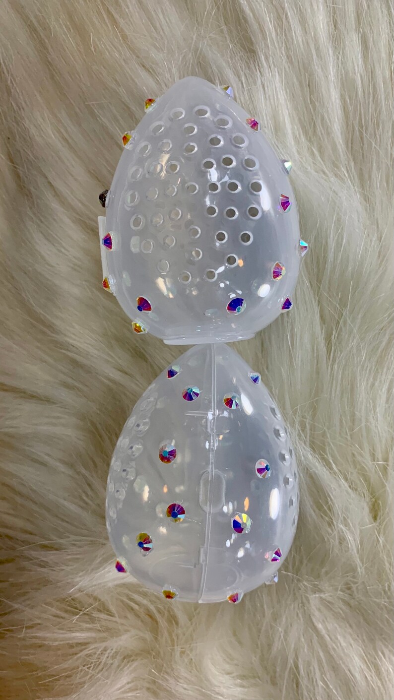 May include: Two clear plastic egg-shaped makeup sponge holders with iridescent rhinestones. The holders are stacked on top of each other.