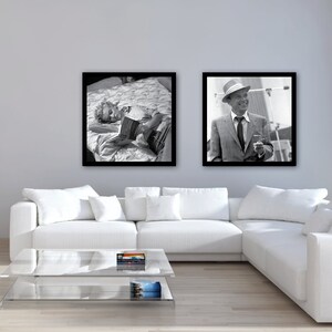 May include: Two black and white framed photographs hanging on a light gray wall above a white sectional sofa. The photo on the left shows a person lying on a bed reading a book. The photo on the right shows a man in a suit and hat holding a cigarette.
