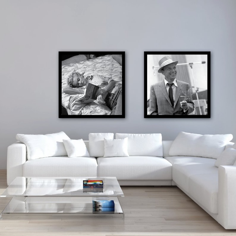 May include: Two black and white framed photographs hanging on a light gray wall above a white sectional sofa. The photo on the left shows a person lying on a bed reading a book. The photo on the right shows a man in a suit and hat holding a cigarette.
