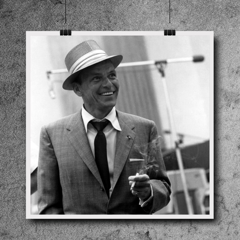 May include: A black and white photo of a man wearing a light-colored fedora, a suit, and a tie. He is holding a cigarette in his right hand and smiling.