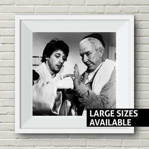 Op de afbeelding: Zwart-wit ingelijste print met twee mannen, waarvan één de hand van de ander verbindt. De print zit in een witte lijst met de tekst "LARGE SIZES AVAILABLE" in een zwarte balk. De afbeelding staat tegen een witte bakstenen muur.