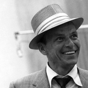 May include: A black and white photo of a man wearing a gray fedora with a white band and a suit jacket. He is smiling.