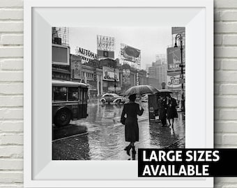 May include: A black and white photograph of a rainy city street scene. A woman with an umbrella walks down the street, passing a bus and other pedestrians. The street is lined with buildings with large signs and advertisements. The photo has the text "LARGE SIZES AVAILABLE" at the bottom.