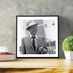 May include: Black and white photo of a man in a suit and hat, holding a cigarette. The man is smiling and looking to the right. The photo is framed in a black frame.