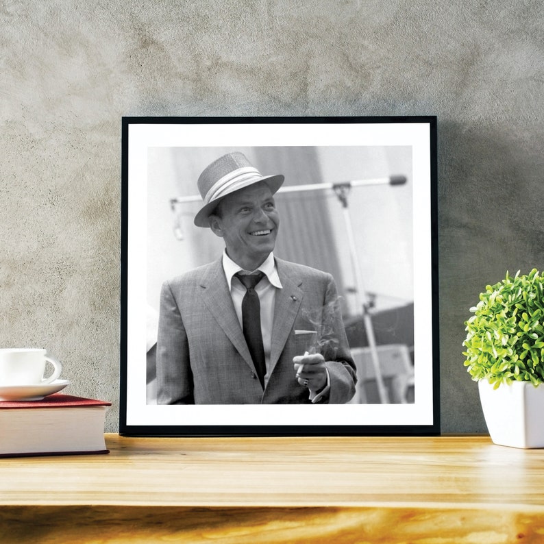 May include: Black and white photo of a man in a suit and hat, holding a cigarette. The man is smiling and looking to the right. The photo is framed in a black frame.