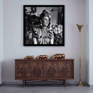 May include: A black and white framed photograph of Frida Kahlo, a Mexican painter, wearing a flower crown and a checkered dress. The photograph is hanging on a white wall above a wooden cabinet with three bronze hippopotamus figurines on top.