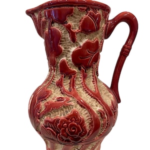 May include: A red ceramic pitcher with a white floral design. The pitcher has a wide mouth and a handle.