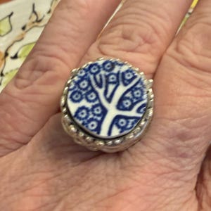 May include: A circular ring featuring a white ceramic inlay with a blue floral tree pattern. The inlay is set within a textured, metallic silver-toned band that has a beaded or granulated edge detail surrounding the central ceramic piece.