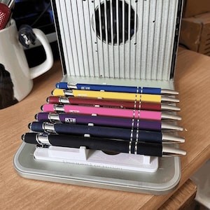 Op de afbeelding: Een verzameling kleurrijke pennen in een metalen etui. De pennen zijn in verschillende kleuren, waaronder blauw, geel, rood, roze, paars en zwart. Elke pen heeft een zilveren punt en een zilveren band. De etui is open en toont de pennen netjes gerangschikt.