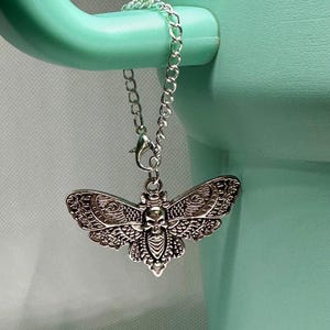May include: A silver-colored moth pendant with a skull design, hanging from a chain. The pendant is attached to a light green mug handle. The moth has detailed wing patterns and a skull in the center.