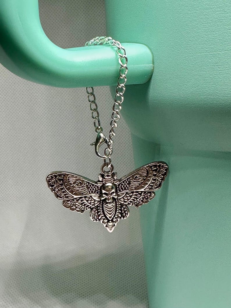 May include: A silver-colored moth pendant with a skull design, hanging from a chain. The pendant is attached to a light green mug handle. The moth has detailed wing patterns and a skull in the center.