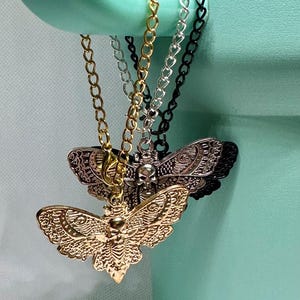 May include: Close-up of several moth pendant necklaces. The pendants are gold, silver, and black, with intricate details. The necklaces hang from a light green object. The moth pendants have a skull design.