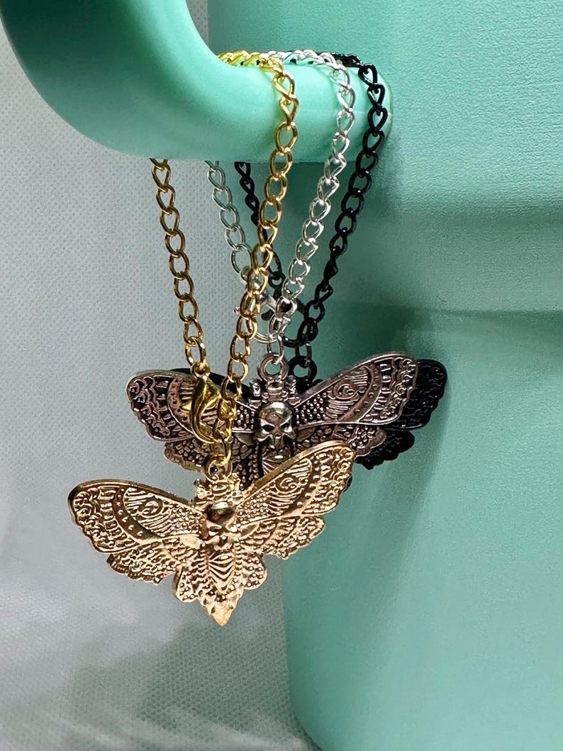 May include: Close-up of several moth pendant necklaces. The pendants are gold, silver, and black, with intricate details. The necklaces hang from a light green object. The moth pendants have a skull design.