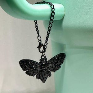 May include: A black metal moth charm with intricate wing detailing, suspended from a delicate chain. The charm is attached to a light green object. The moth's design is highly detailed, showcasing its wings and body. The chain is made of small, heart-shaped links.