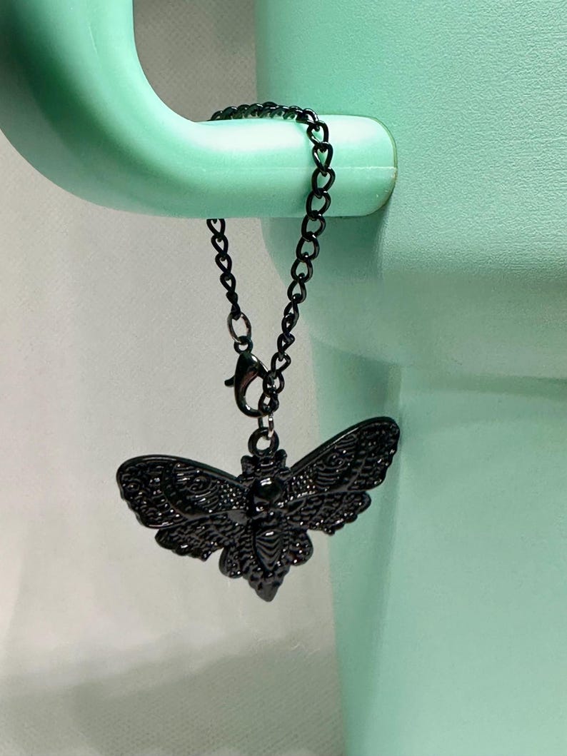 May include: A black metal moth charm with intricate wing detailing, suspended from a delicate chain. The charm is attached to a light green object. The moth's design is highly detailed, showcasing its wings and body. The chain is made of small, heart-shaped links.