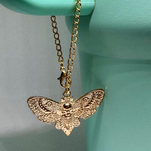 May include: A gold-colored moth charm pendant with intricate wing detailing hangs from a delicate gold chain. The charm is suspended against a light teal background, creating a striking contrast. The chain is attached to a teal-colored object.