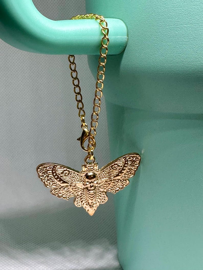 May include: A gold-colored moth charm pendant with intricate wing detailing hangs from a delicate gold chain. The charm is suspended against a light teal background, creating a striking contrast. The chain is attached to a teal-colored object.
