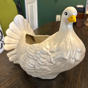 May include: A white ceramic turkey-shaped planter with a pearlescent finish. The bird features a fanned tail, textured wing details, a bright yellow beak, and painted black eyes with eyelashes. The hollow center provides space for small items.