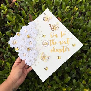 May include: A white graduation cap decorated with white flowers, gold butterflies, and pearls. The cap has the text "On to the next chapter" in gold script. The cap is held against a background of green leaves.