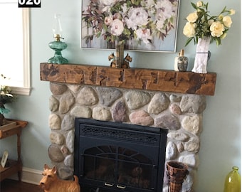 Op de afbeelding: Een stenen open haard met een donkere metalen vuurhaard, bekroond met een houten mantel. De mantel bevat een groene glazen lamp, een schilderij van witte bloemen en decoratieve items. Een houten hondenbeeldje staat op de haard.