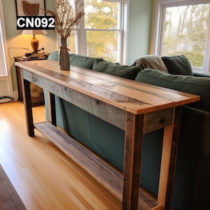 May include: A long, rustic wooden console table with a shelf. The table top is made of reclaimed wood with a variety of colours and textures. The table is in a living room setting, behind a green sofa.