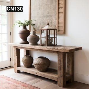 May include: A rustic wooden console table with a lower shelf. The table is decorated with three pottery vases and a glass lantern. The table is in front of a white wall and a window.