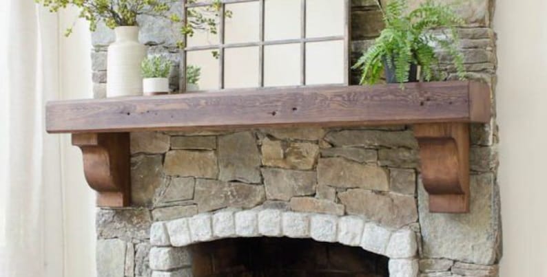 May include: A rustic wooden mantel shelf with a stone fireplace below. The mantel is supported by two wooden brackets. The fireplace has a stone archway and a black metal grate.