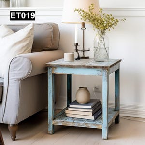 May include: A light blue end table with a distressed wood finish and a lower shelf. The table has a vase with greenery, a small ceramic vase, and a stack of books on the shelf. The table is labelled "ET019".