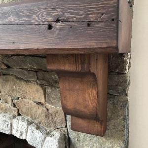 May include: A rustic wooden mantel shelf with a dark brown stain, mounted above a stone fireplace. The shelf has a simple, rectangular design with a slightly curved edge on the front. The shelf is supported by two brackets, which are also made of wood and have a dark brown stain.
