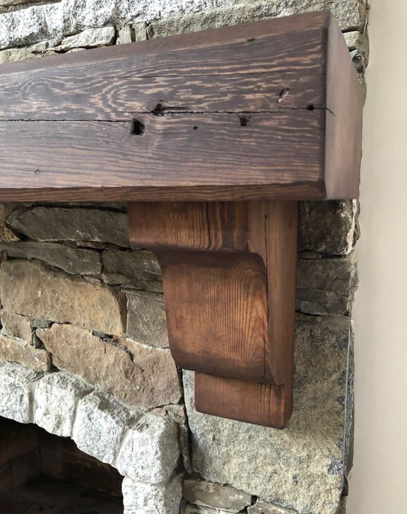 May include: A rustic wooden mantel shelf with a dark brown stain, mounted above a stone fireplace. The shelf has a simple, rectangular design with a slightly curved edge on the front. The shelf is supported by two brackets, which are also made of wood and have a dark brown stain.