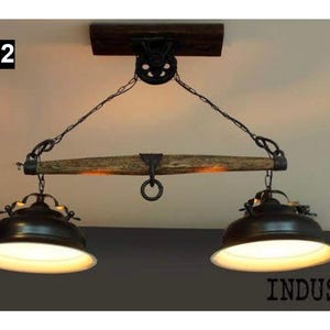 May include: Rustic-style ceiling light fixture with two lamps. The fixture features a dark brown wooden beam, black metal chains, and two black lampshades with white interiors. The text "INDUS3'L" is visible at the bottom.