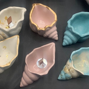 May include: Six seashell-shaped ceramic planters in various colors, including white, pink, and blue. Each planter has a gold rim and is decorated with gold flecks. The planters are perfect for small plants or succulents.