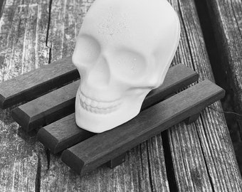 May include: A white ceramic skull with a toothy grin sits on a wooden soap dish. The skull has a textured surface and is slightly tilted to the side.