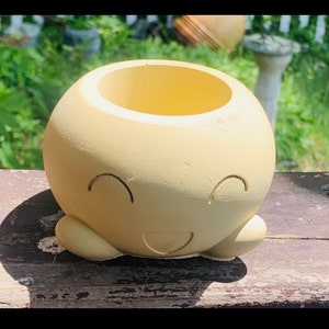 May include: A light yellow ceramic planter shaped like a smiling face with two small arms. The planter has a round opening at the top for planting.
