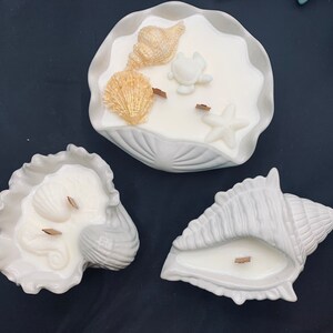 May include: Three white ceramic seashell candle holders. The largest seashell has a white wax candle with gold seashell and starfish decorations. The other two seashells have white wax candles.