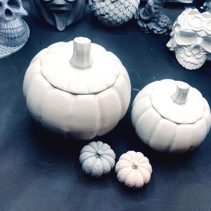 May include: Two white ceramic pumpkin-shaped containers with lids, one larger than the other.  Two smaller decorative pumpkins, one gray and one white, are in the foreground.