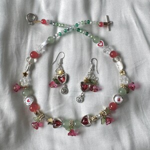 May include: A green and red beaded necklace with heart-shaped charms and matching earrings. The necklace has a silver clasp and the earrings have silver hooks.