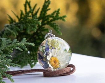 May include: A clear, spherical pendant necklace containing a preserved daisy and other small flowers. The pendant is suspended from a brown leather cord. Green foliage is in the background, with a white surface in the foreground.