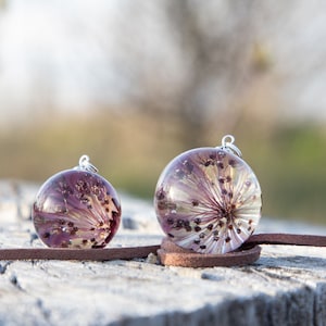 Puede incluir: Dos colgantes redondos de resina con flores secas encapsuladas en resina transparente. Los colgantes están en un cordón de cuero marrón y están colgando de un trozo de madera desgastada.