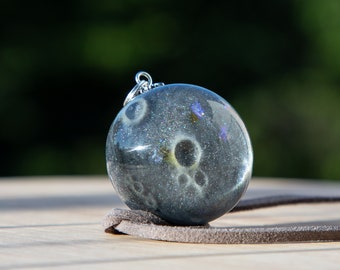 May include: A round, gray pendant with a silver bail. The pendant is filled with a clear resin that contains small, dried flowers and other botanical elements. The pendant is hanging from a brown leather cord.