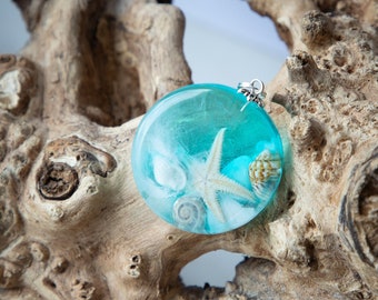 May include: A round pendant with a blue resin background and a white starfish and seashells inside. The pendant is on a piece of driftwood.