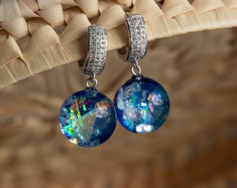 May include: A pair of silver hoop earrings with a sparkling, clear stone design. Each earring features a round, dark blue pendant with iridescent, colorful accents. The earrings are displayed against a woven, light brown background.