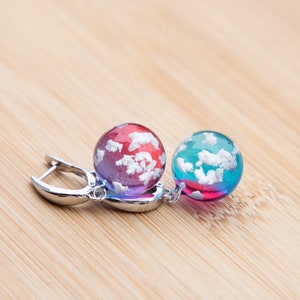 May include: A pair of earrings with silver hoops and round, colorful glass beads. The beads are filled with white swirls, resembling clouds. One bead is red and blue, the other is blue and green.