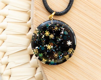 May include: A round, black pendant necklace with a gold-colored bail and a black cord. The pendant is filled with glitter and snowflake shapes in gold, silver, and teal. The necklace rests on a light brown wooden surface.