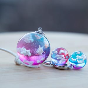 May include: A set of three resin jewelry pieces featuring a cloudy sky with pink, blue, and red hues. The largest piece is a pendant with a silver chain, and the two smaller pieces are earrings.