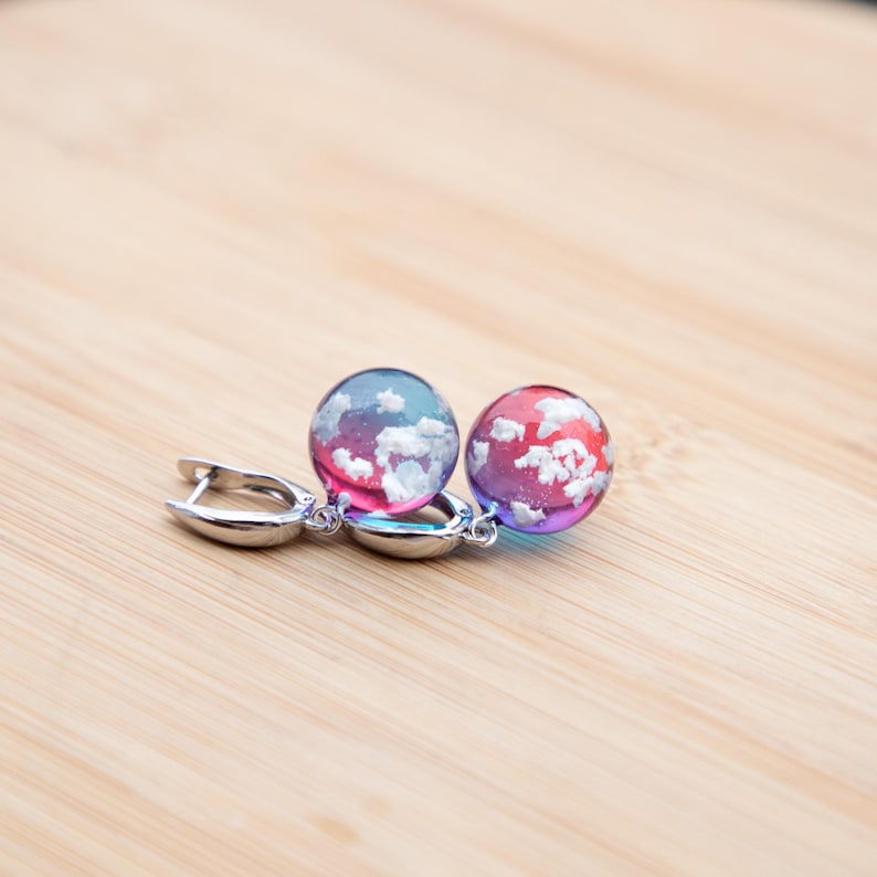 May include: A pair of silver hoop earrings with a pink and blue gradient resin sphere with white flecks, resembling clouds, hanging from each hoop.