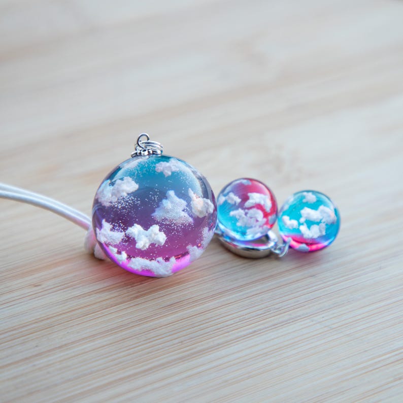 May include: Three resin ornaments with a cloudy sky design. The largest ornament is a pendant with a silver bail. The smaller ornaments are blue and pink with white clouds.