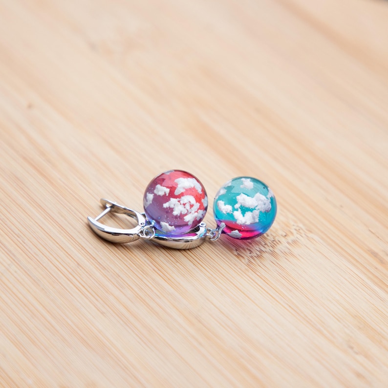 May include: A pair of earrings with silver hoops and round, colorful glass beads. The beads are filled with white swirls, resembling clouds. One bead is red and blue, the other is blue and green.
